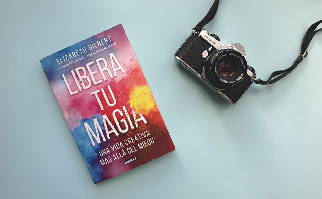 Portada del libro de autoayuda Libera tu magia de la autora Elizabeth Gilbert en un fondo azul pastel al lado de una cámara pentax análoga