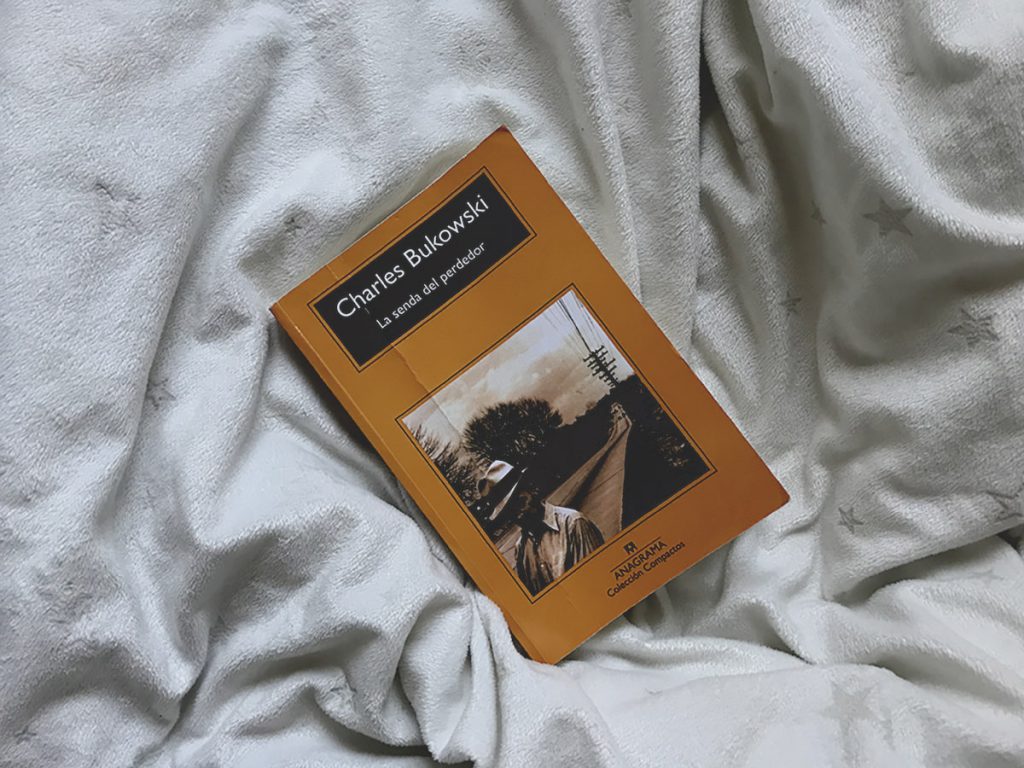 Libro La senda del perdedor de escritor Charles Bukowski sobre una cobija blanca.