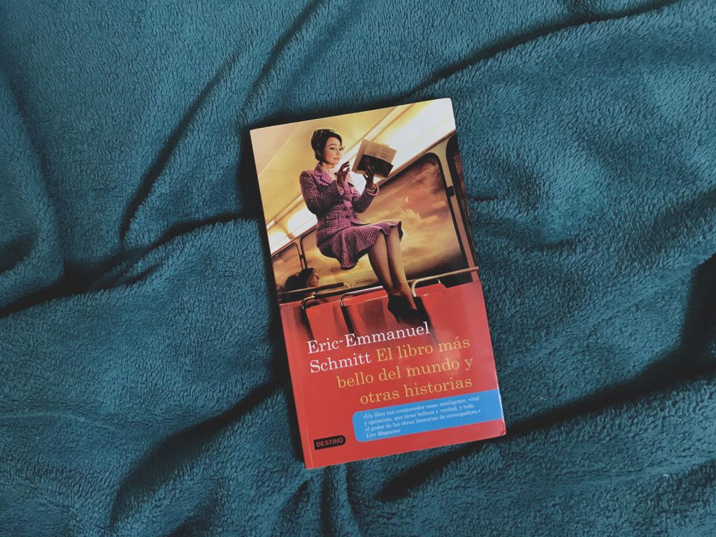 El libro titulado "El libro más bello del mundo y otras historias" escrito por Eric-Emmanuel Schmitt está sobre una cobiza azul. La portada es una señora leyendo en un autobús mientras ella está flotando sobre los asientos. 