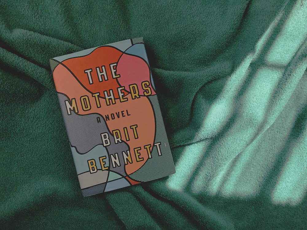 Libro novela The Mothers de la escritora Brit Bennett sobre cobija verde. La portada es muy colorida, es el perfil de la cara de una mujer pero muy abstracto, sin ojos ni detalles, solo líneas en la piel y bloque de colores naranja y rojo en el cabello. 